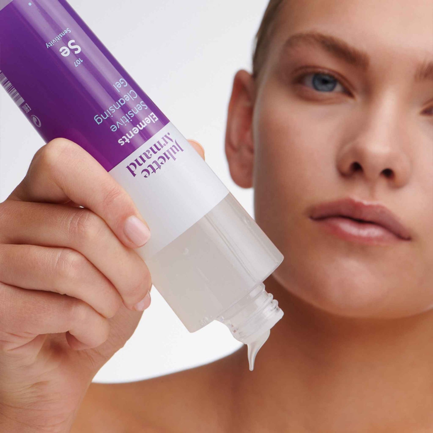 Hand holding a Juliette Armand Elements Sensitive Cleansing Gel 210mL  near a person's face on a white background